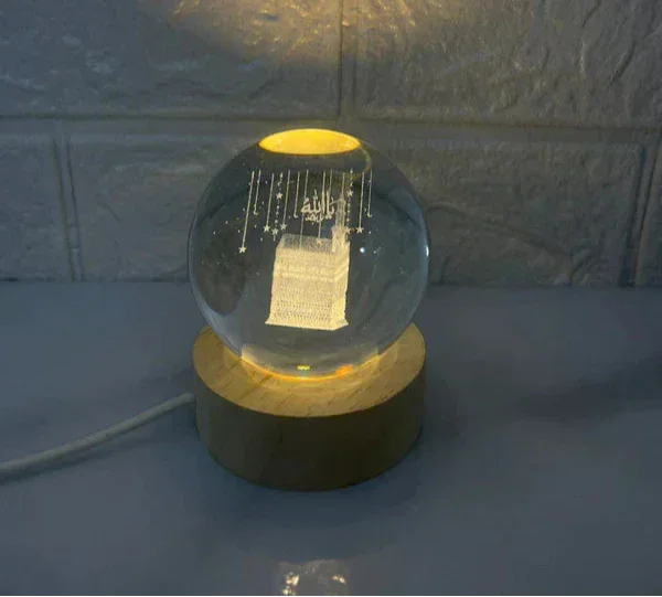 Illuminated crystal globe lamp with 3D Mecca Kaaba design on wooden base, glowing in dim light
