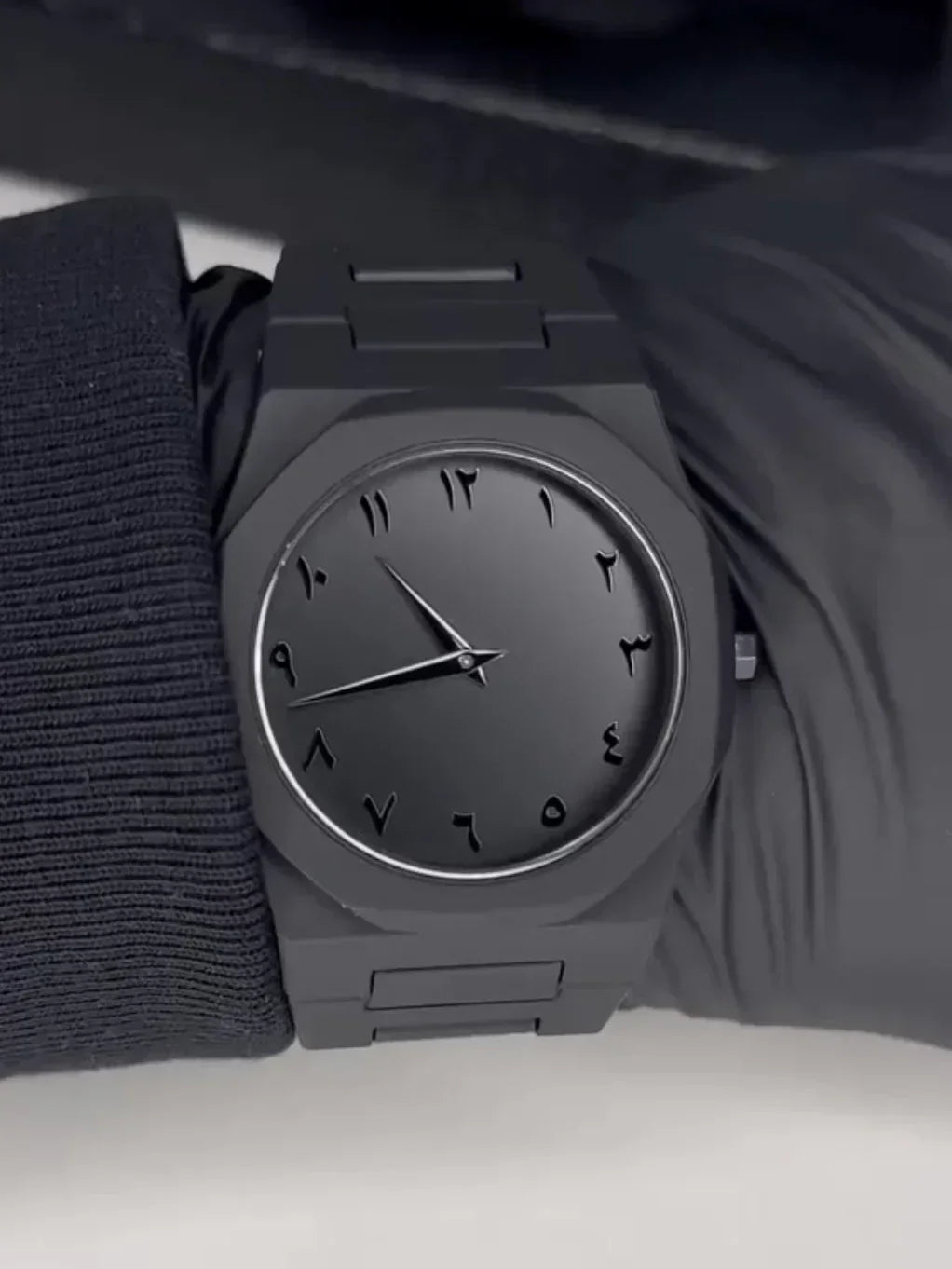 Black wristwatch with Arabic numerals and matte black metal band on a gloved wrist