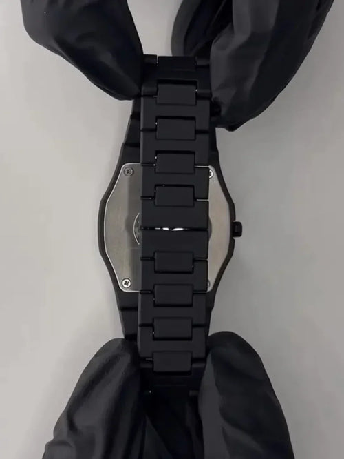 Black metal wristwatch band with clasp, held by gloved hands on a white background